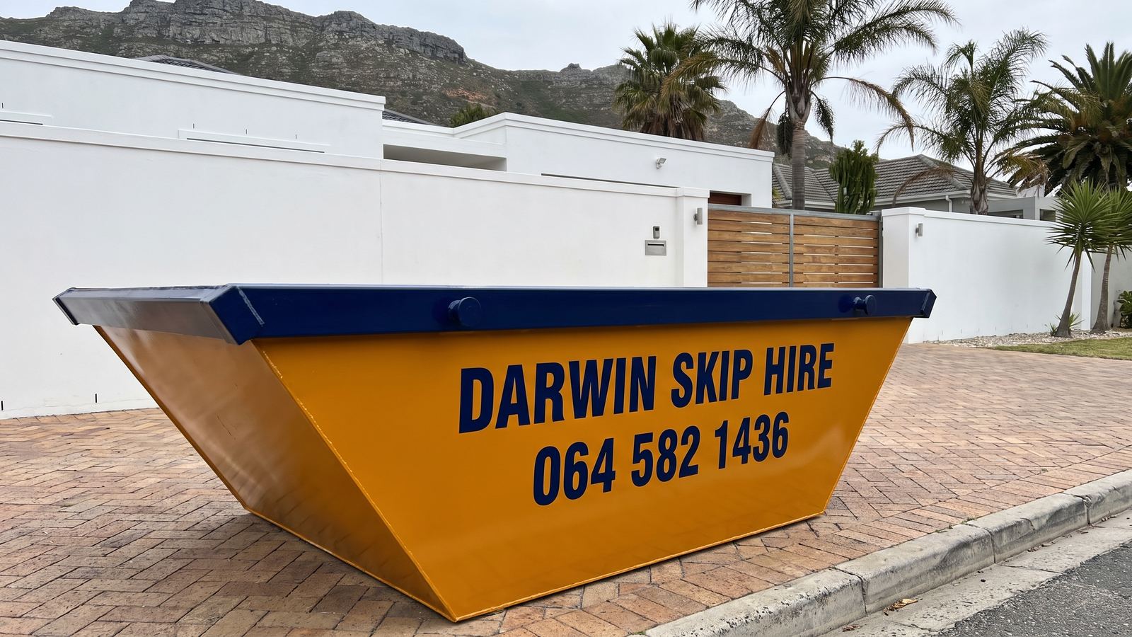 Amber Darwin mini skip on a brick driveway in front of a white villa with palms and a mountain ridge.