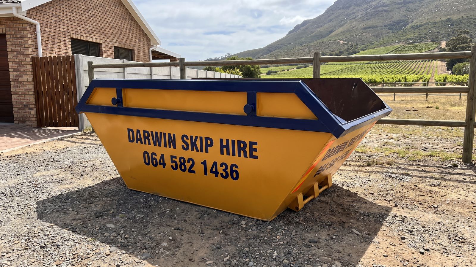 Amber Darwin mini skip on a gravel yard beside a post-and-rail fence with vineyards behind.