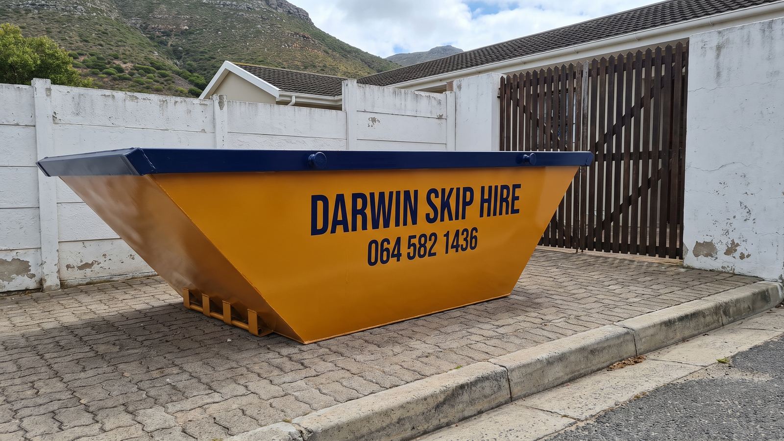 Amber Darwin mini skip on paving beside a white wall and wooden gate below a mountain slope.