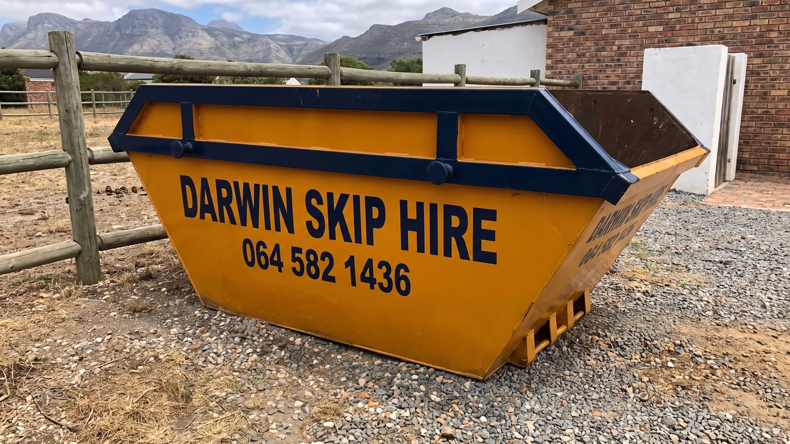 Amber Darwin mini skip on a gravel yard beside a post-and-rail paddock fence with mountains behind.