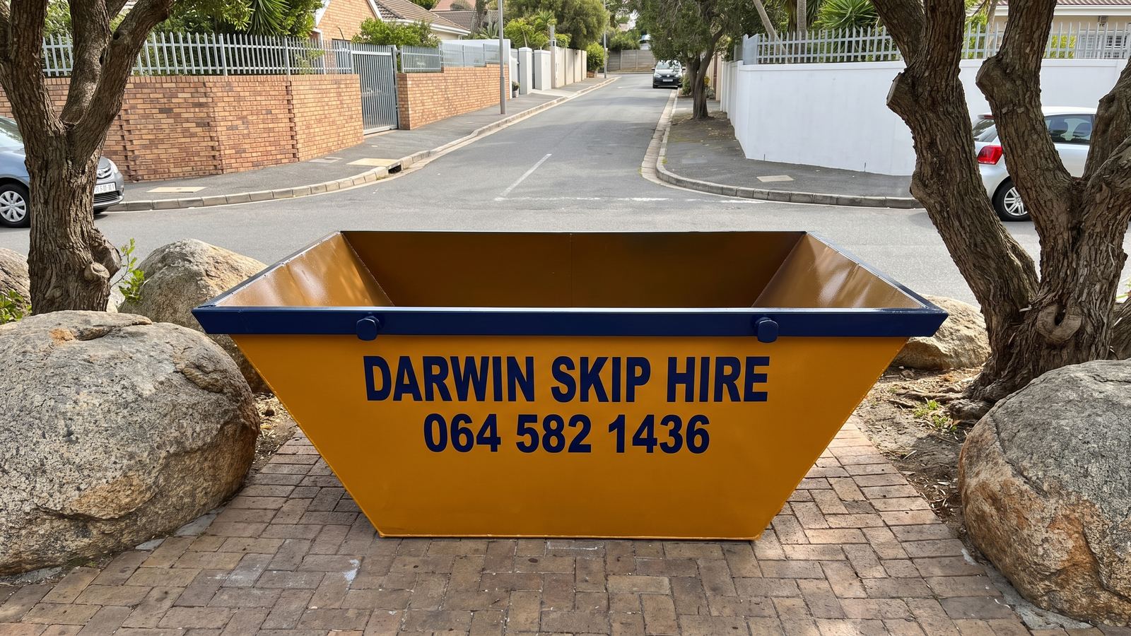 Amber Darwin mini skip on a paved driveway between granite boulders and milkwood trees.