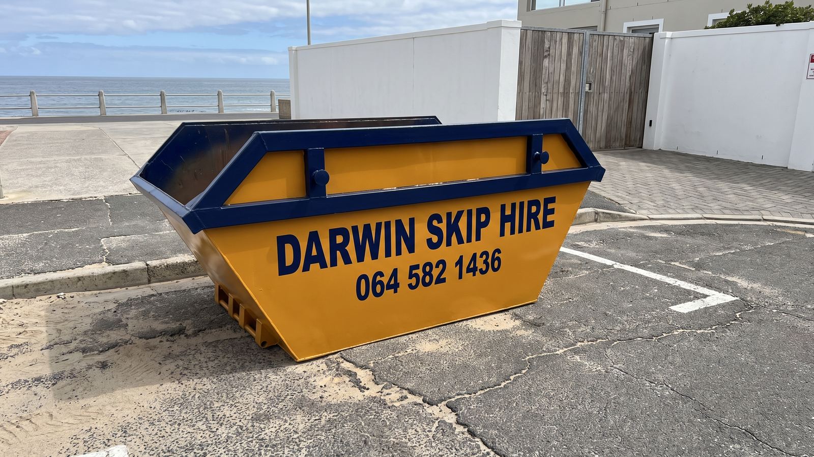 Amber Darwin mini skip in a ground-level parking area near a sea promenade.