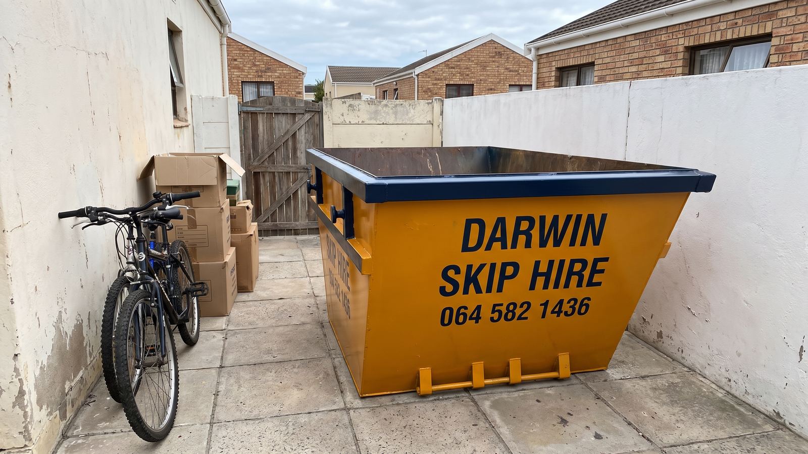 Amber Darwin mini skip in a narrow paved yard beside a bicycle and moving boxes.