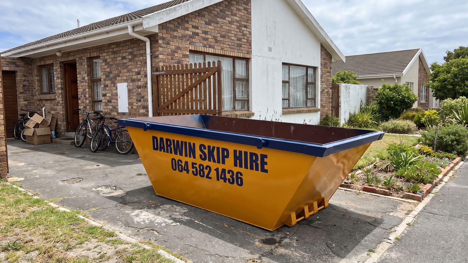 Amber Darwin mini skip on the driveway of a brick house with bicycles and boxes by the door.
