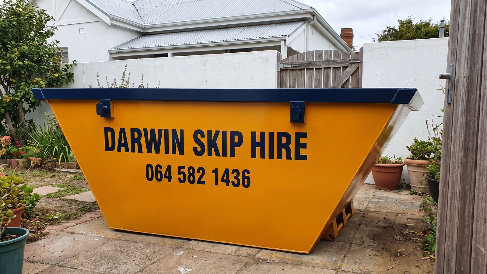 Amber Darwin mini skip in a small paved yard with pot plants beside a wooden gate.