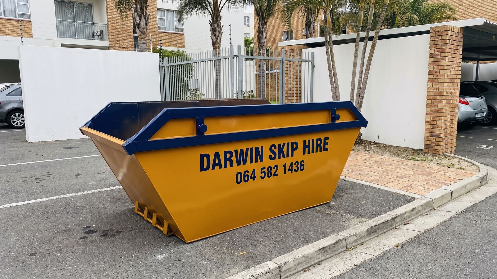 Amber Darwin mini skip in a ground-floor parking bay of an apartment block with palm trees.