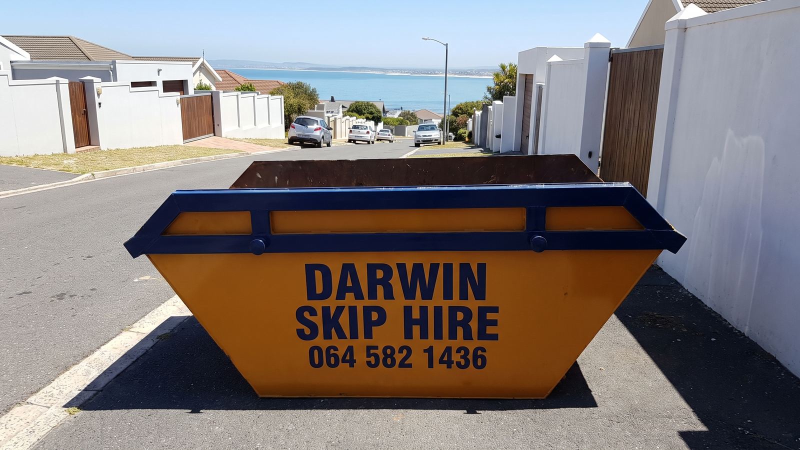 Amber Darwin mini skip standing in a steep residential street with a bay in the distance.